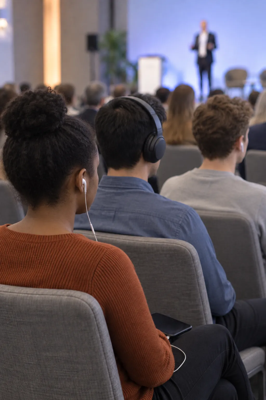 In a congress hall three people in one row listen side by side, through a wired earphone, through over ear headphones and through a white earphone; all eyes are on the platform.